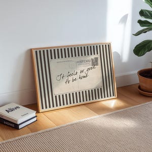 Vintage postcard-style wall art with handwritten script reading “It feels so good to be home,” set on a neutral striped background in a cozy sunlit interior.