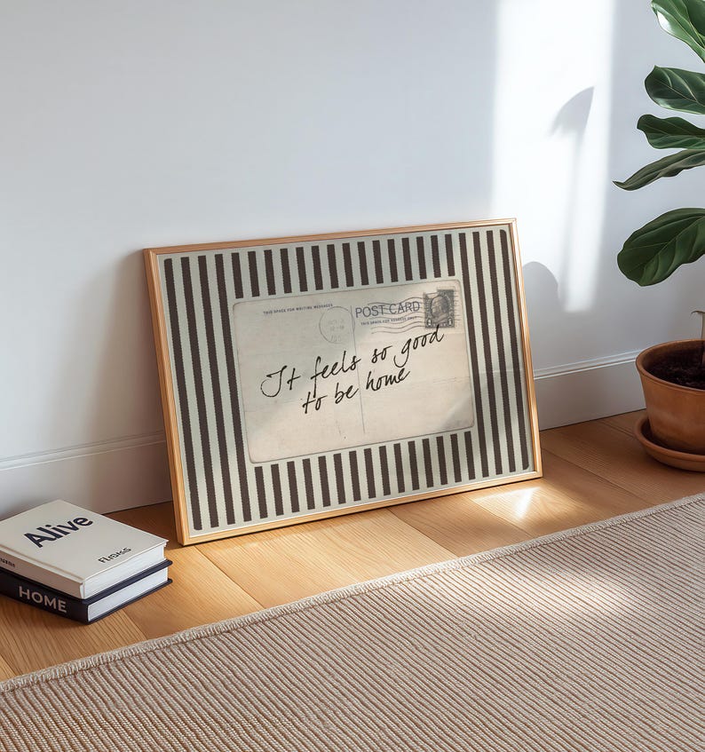 Vintage postcard-style wall art with handwritten script reading “It feels so good to be home,” set on a neutral striped background in a cozy sunlit interior.