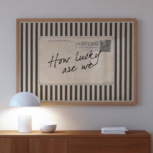 Framed vintage-style digital art featuring a sepia-toned postcard with the handwritten phrase “How lucky are we.” The postcard is set against a background of vertical brown stripes, adding a graphic yet nostalgic look. Perfect for gallery walls, vintage-inspired interiors, or thoughtful gifts.