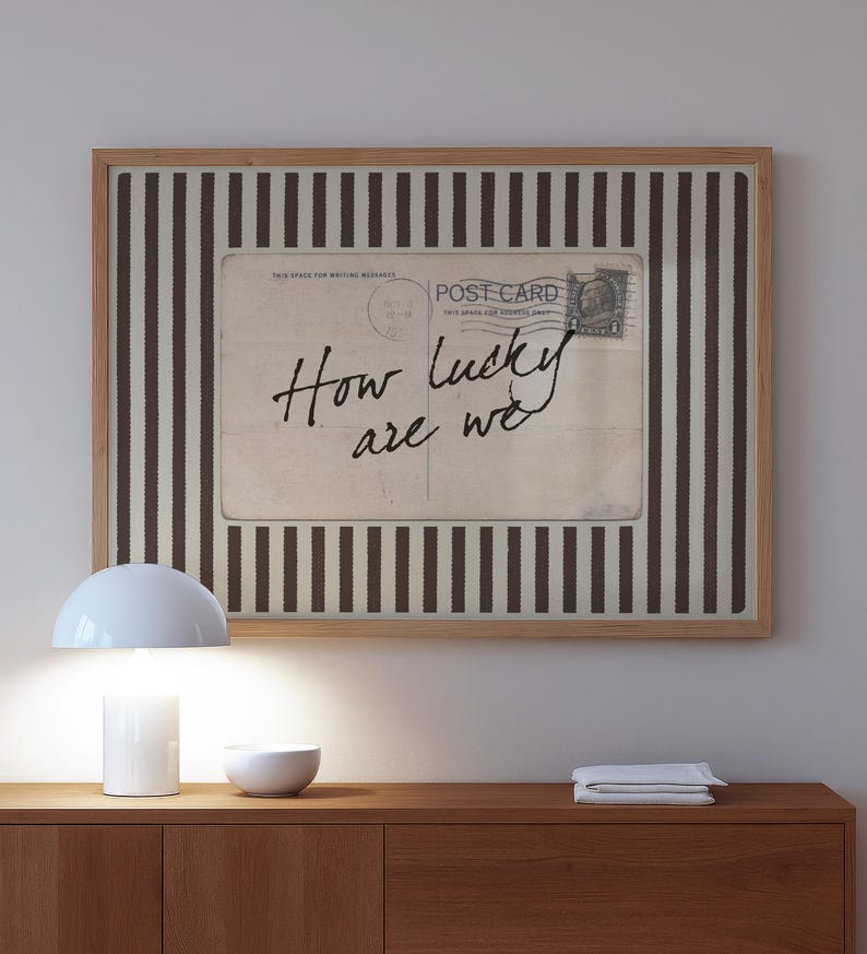 Framed vintage-style digital art featuring a sepia-toned postcard with the handwritten phrase “How lucky are we.” The postcard is set against a background of vertical brown stripes, adding a graphic yet nostalgic look. Perfect for gallery walls, vintage-inspired interiors, or thoughtful gifts.