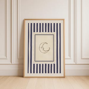 Vintage-inspired printable wall art featuring a hand-drawn crescent moon illustration with a softly smiling face, centered within a muted parchment-style frame. The moon is surrounded by layered, scalloped-edge borders and encased in deep navy vertical stripes, adding a celestial yet refined aesthetic. Styled in a minimalist, quiet luxury interior with warm wood flooring and decorative wall paneling, this digital download evokes dreamy, moody nighttime vibes.