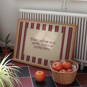 Op de afbeelding: Een ingelijst kunstwerk met een licht houten frame, met een crèmekleurig midden met de tekst "Your custom text, quote, lyrics or saying here." Het kunstwerk heeft een rood-witte gestreepte rand. Een mand met appels en een enkele appel staan op de voorgrond.