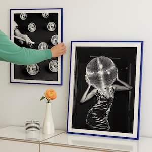 May include: Two framed black and white art prints with blue borders. One print features disco balls, the other a person with a disco ball head. A green-sleeved arm is holding one of the prints. A white vase with a single orange flower is on a white surface.