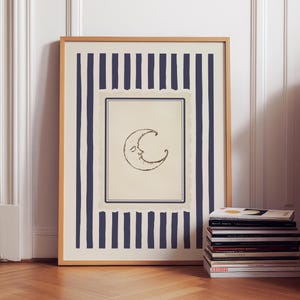 Vintage-inspired printable wall art featuring a hand-drawn crescent moon illustration with a softly smiling face, centered within a muted parchment-style frame. The moon is surrounded by layered, scalloped-edge borders and encased in deep navy vertical stripes, adding a celestial yet refined aesthetic. Styled in a minimalist, quiet luxury interior with warm wood flooring and decorative wall paneling, this digital download evokes dreamy, moody nighttime vibes.