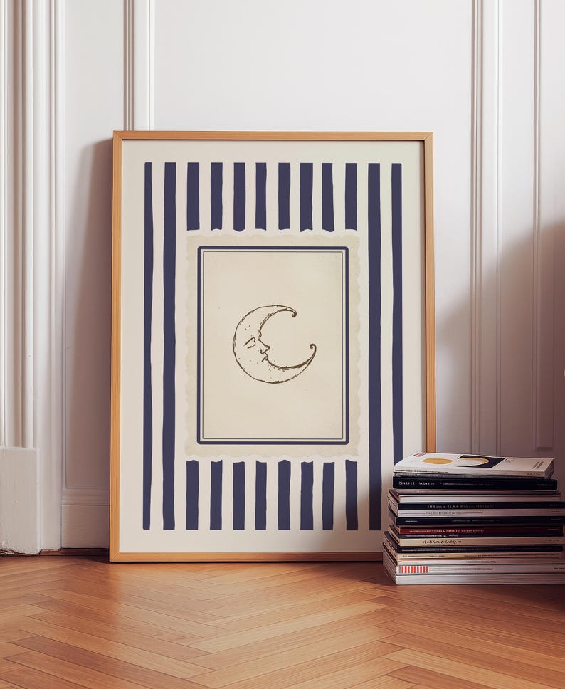 Vintage-inspired printable wall art featuring a hand-drawn crescent moon illustration with a softly smiling face, centered within a muted parchment-style frame. The moon is surrounded by layered, scalloped-edge borders and encased in deep navy vertical stripes, adding a celestial yet refined aesthetic. Styled in a minimalist, quiet luxury interior with warm wood flooring and decorative wall paneling, this digital download evokes dreamy, moody nighttime vibes.