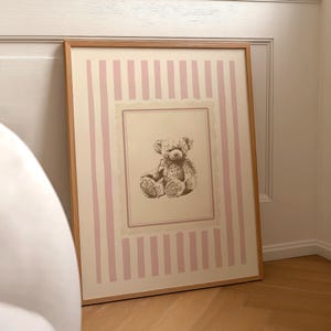 May include: A framed print featuring a vintage teddy bear illustration. The artwork has a light pink and cream striped background with a scalloped border. The frame is made of light-colored wood.