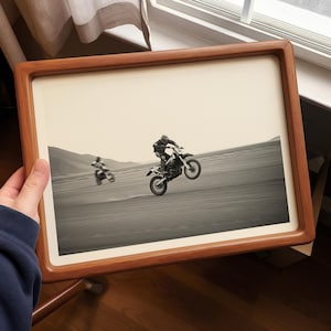 Puede incluir: Impresión enmarcada en blanco y negro de dos motos de cross corriendo por un paisaje desértico. Una moto está en medio de un caballito, la otra está más lejos. El marco es de color marrón cálido.