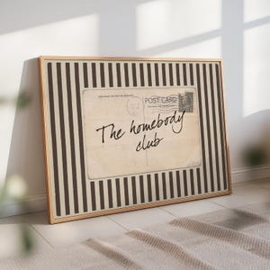 Vintage postcard-style wall art reading “The homebody club,” displayed on a neutral striped background in a softly lit, cozy interior.