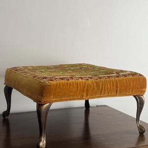 May include: An antique footstool with a square, mustard-yellow velvet cushion. The cushion is adorned with a floral tapestry. The footstool has four curved, bronze-colored legs, adding to its vintage charm. The footstool is a decorative furniture piece.