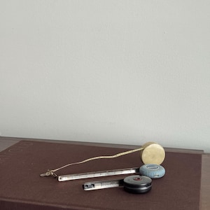 Może przedstawiać: Kolekcja starych taśm mierniczych w r&oacute;żnych stylach. Obejmuje beżową, okrągłą taśmę mierniczą, niebiesko-srebrną taśmę mierniczą oraz czarno-srebrną taśmę mierniczą. Taśmy leżą na brązowej książce.