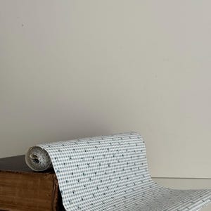 May include: A roll of white fabric with a blue floral pattern, unrolled on top of a vintage brown book. The fabric has a gingham-like pattern with small blue floral accents. The book has a worn, aged appearance.