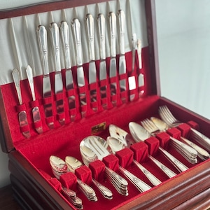 May include: A wooden silverware chest with a red velvet interior, holding a complete set of silver-colored flatware. The set includes knives, forks, spoons, and serving utensils, all neatly arranged in individual slots. The chest is open, revealing the contents.