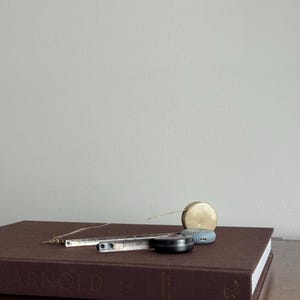 Może przedstawiać: Brązowa książka w twardej oprawie z wytłoczonym słowem "ARNOLD" na okładce. Metalowa linijka, czarna miarka, niebieska miarka i złota miarka znajdują się na książce.