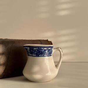 May include: A small, white ceramic creamer with a blue floral pattern around the rim. The creamer has a curved handle and a rounded body. Beside it is a brown leather-bound book.