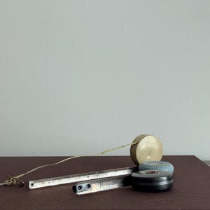 Może przedstawiać: Kolekcja zabytkowych narzędzi pomiarowych, w tym metalowa linijka, mała beżowa miarka i dwa okrągłe urządzenia pomiarowe. Narzędzia są ułożone na brązowej powierzchni, co sugeruje skupienie się na rzemiośle lub historycznych narzędziach.