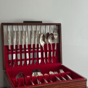 May include: A wooden storage chest with a red velvet interior containing a set of silver-toned flatware. The top section holds upright knives, spoons, and forks with floral embossed handles, while the bottom drawer contains additional utensils.
