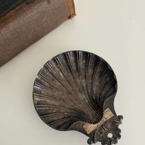 May include: A vintage, silver-toned seashell-shaped dish with a decorative floral accent. The dish has a textured, ridged surface and a small hole near the floral design. A worn, brown leather-bound book is partially visible in the background.