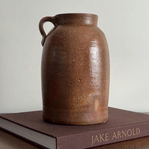 May include: A brown ceramic jug with a cylindrical body, a short neck, and a single curved handle on the side. The surface of the jug has a textured, speckled appearance with a glossy finish, showing variations in the brown color throughout the piece.