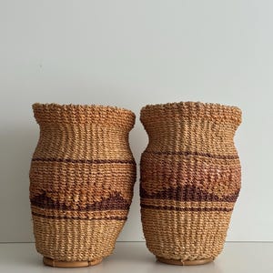 May include: Two woven baskets with a natural, light brown color and dark brown horizontal stripes. The baskets have a rounded shape with a slightly flared top and sit on a circular base. The texture of the baskets is rough and organic.