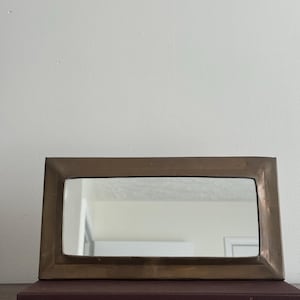 May include: A rectangular bronze-colored mirror with a simple frame. The mirror is resting on a dark purple book with the title "JAKE ARNOLD" in gold lettering. The background is a plain white wall.
