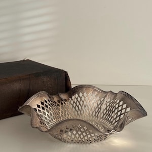May include: A silver-colored, openwork bowl with a ruffled edge. The bowl has a diamond-shaped pattern throughout its design. A dark brown book is partially visible in the background.