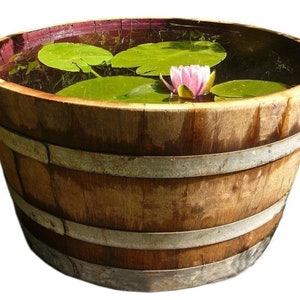 May include: A wooden barrel planter with metal bands, filled with water, lily pads, and a pink water lily. The barrel has a natural wood grain finish and is a decorative outdoor garden element.