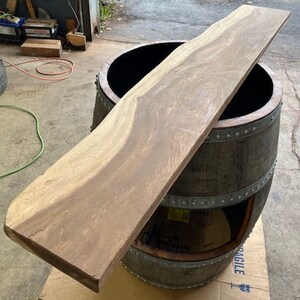 May include: A light brown wooden plank rests on top of a dark wooden barrel with metal bands. The plank has a natural edge and visible wood grain. The barrel is open at the top and has a dark interior.