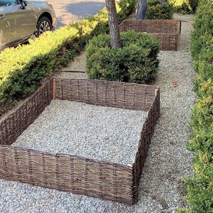 May include: Two square wicker garden beds filled with gravel. The beds are surrounded by green bushes and a gravel path.