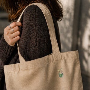 May include: Beige canvas tote bag with a small, colorful plant illustration. The bag is carried over the shoulder, showcasing its simple design and natural color. The plant illustration features green leaves and a yellow sprout.