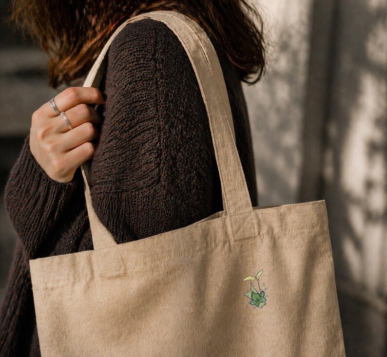 May include: Beige canvas tote bag with a small, colorful plant illustration. The bag is carried over the shoulder, showcasing its simple design and natural color. The plant illustration features green leaves and a yellow sprout.
