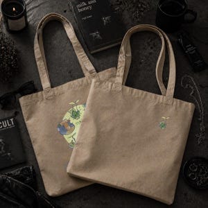 May include: Two beige canvas tote bags with long handles. One bag features a colorful graphic print, while the other has a small embroidered design. The bags are on a dark surface, surrounded by accessories.
