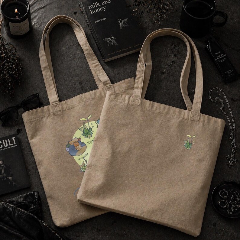 May include: Two beige canvas tote bags with long handles. One bag features a colorful graphic print, while the other has a small embroidered design. The bags are on a dark surface, surrounded by accessories.