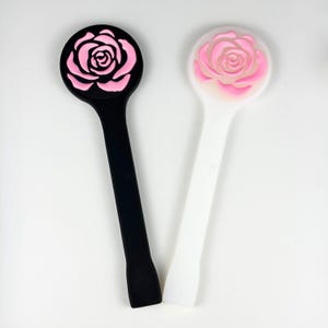 May include: Two long-handled cosmetic spatulas, one black and one white, each featuring a circular top end decorated with a raised, three-dimensional pink rose design. The tools have a smooth, flat handle and a rounded, wide head for application.