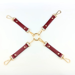 May include: A burgundy leather harness with gold-colored metal hardware. The harness features four straps connected to a central ring, each ending in a clasp. The straps are secured with small gold rivets.