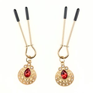 May include: A pair of gold-coloured nipple clamps with black tips. Each clamp has a decorative pendant with a red teardrop-shaped gem surrounded by small white pearls and ornate gold filigree.
