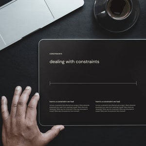 May include: A person is using a stylus to write on a black tablet with the text "dealing with constraints" on the screen. The tablet is on a black surface with a laptop and a cup of coffee.