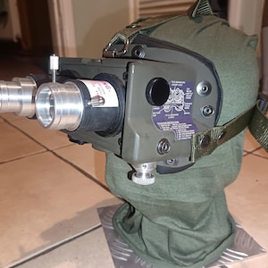 May include: An olive green night vision device with silver lenses and a black rubber casing. The device is mounted on a green fabric head covering with adjustable straps. A label with text is visible on the device.