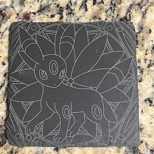 May include: A black slate coaster with a white line drawing of a Pokemon character, a dark-type Eeveelution, with a mandala-like design in the background.