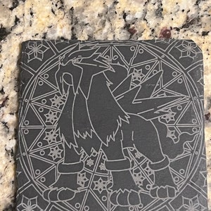 May include: Black and white engraved image of a Pokemon character on a black slate coaster. The character is standing in the center of a circular design.