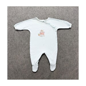 May include: A light blue long-sleeved footed sleeper with a diagonal snap closure at the neckline. The center of the chest features a small embroidered graphic of a puppy sitting in a red wagon with a colorful ball beside it.
