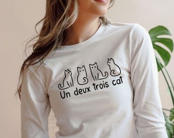 Pode incluir: Camiseta branca de manga comprida com um gráfico preto de quatro gatos sentados em fila. O texto "Un deux trois cat" está impresso abaixo dos gatos.