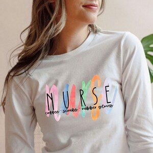 May include: White long-sleeve shirt with a colorful watercolor design that says "NURSE" with the words "coffee, scrubs, rubber gloves" underneath.