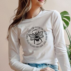 May include: White long-sleeve shirt with a black bee graphic and the text "YOU'RE-HUMBLE-KIND-POSITIVE-FUN-CONFIDENT-BRAVE-SMART-HAPPY-WILD-CREATIVE-JOYFUL-YOURSELF".
