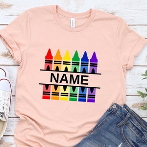 May include: A pink t-shirt with a design of a row of colorful crayons with the word "NAME" in black text in the middle.