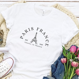 May include: White t-shirt with a black graphic of the Eiffel Tower and the text "Paris France Est 1928 Travel in France".
