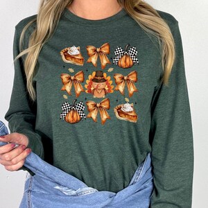 May include: A green long-sleeved t-shirt with a Thanksgiving-themed design. The design features a turkey, pumpkins, pie, and bows in shades of orange, brown, and white.