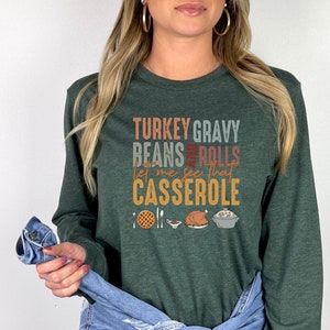 May include: A green long-sleeved t-shirt with a Thanksgiving-themed graphic that reads "Turkey Gravy Beans Rolls Let me see that Casserole". The graphic includes illustrations of a casserole dish, a turkey, a side dish, and a pie.