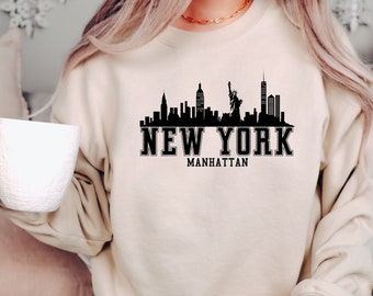 Könnte beinhalten: Ein beiger Pullover mit einem schwarzen Aufdruck der Skyline von New York City und dem Text "NEW YORK MANHATTAN".