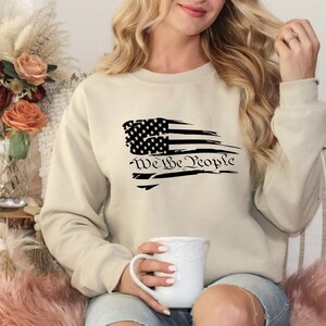 May include: A beige sweatshirt with a distressed American flag design and the text "We the People" in black.