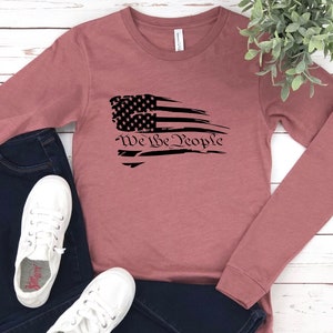 May include: A long-sleeved, mauve t-shirt with a distressed black and white American flag graphic and the text "We the People".